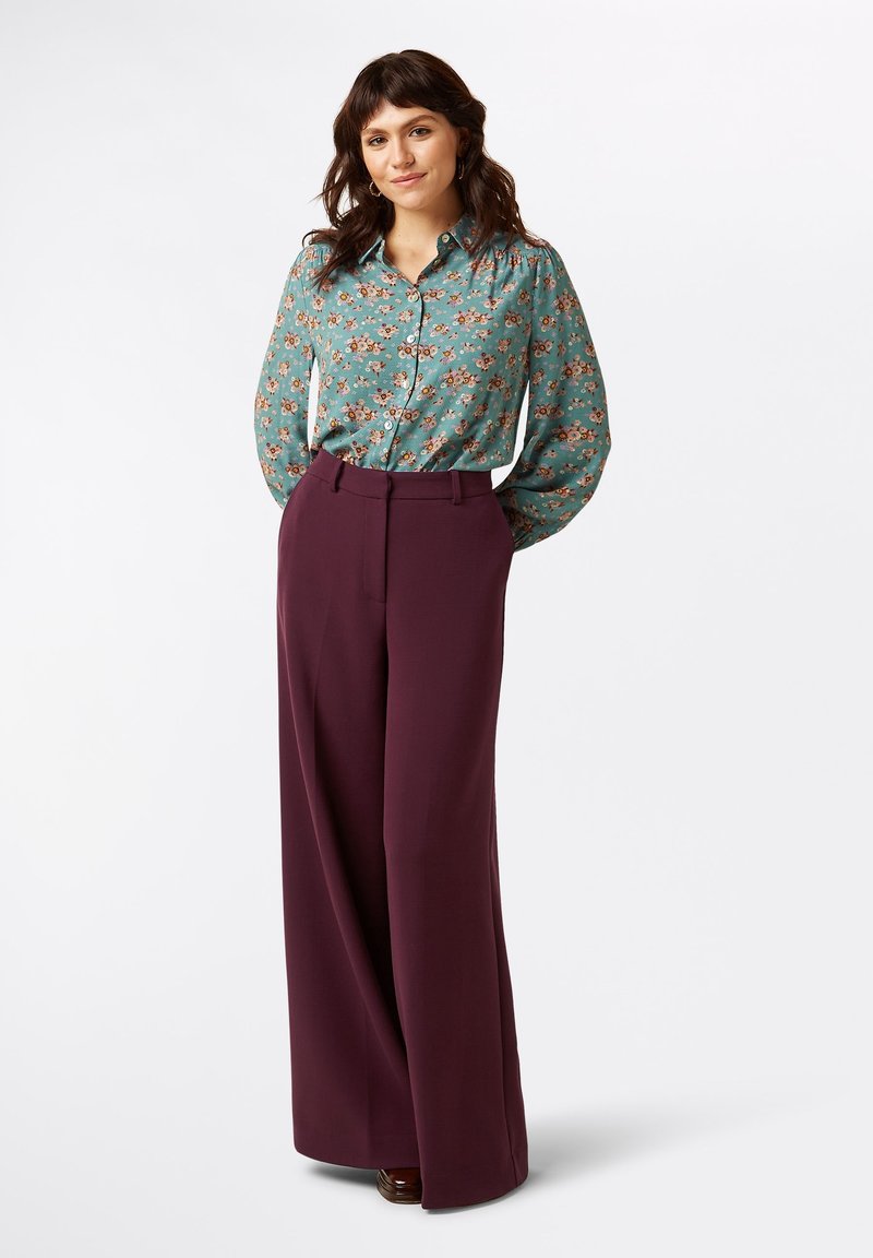 Bluse in Petrol mit floralem Muster und Knopfleiste, kombiniert mit hoch taillierten, weit geschnittenen bordeauxfarbenen Hosen, die eine lockere Passform und eine glatte Textur bieten.