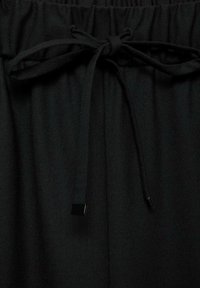 Zwarte elastische taillebandbroek met een striksluiting, voorzien van een platte vierkante metalen accent op de strik en een getextureerd stoffen oppervlak.