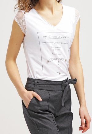 Femme portant un T-shirt blanc à manches en dentelle avec un texte en français et un pantalon gris taille haute noué d'un nœud à la taille.
