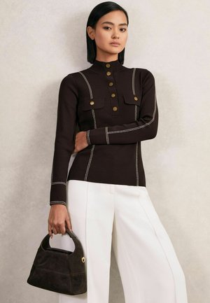Donna che indossa un top a maniche lunghe nero con bottoni e cuciture bianche, pantaloni bianchi, mentre tiene una borsa nera con manico arrotondato.