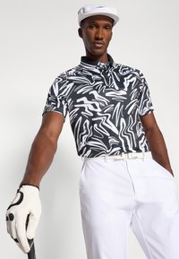 Polo preto e branco com padrão abstrato e colarinho, combinado com calças brancas e um boné branco. Apresenta uma luva de aderência e um taco de golfe.