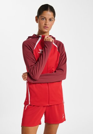 Vrouw met donker haar die een rode sportjas met kastanjebruine mouwen en bijpassende rode shorts draagt, staand met gekruiste armen.