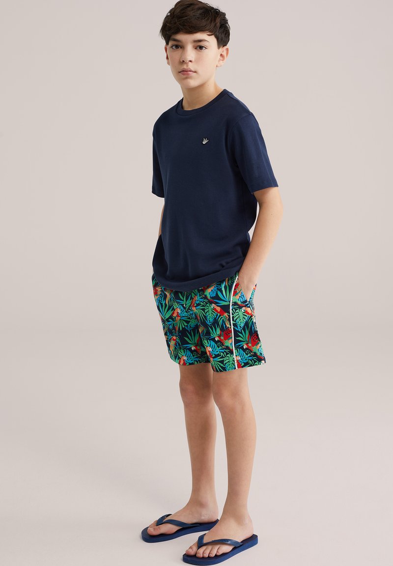 Granatowy bawełniany t-shirt z krótkim rękawem; wzorzyste szorty kąpielowe z zielonymi liśćmi i czerwonymi ptakami; niebieskie klapki. Model stoi z rękami w kieszeniach.