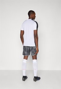 Herre atletisk outfit bestående af en hvid t-shirt med korte ærmer, mønstrede sorte shorts, hvide knæstrømper med røde bogstaver, og sorte fodboldstøvler.