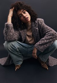 Grau gepunkteter Leopardencape, beiger Rollkragenpullover, hellblaue weite Jeans und spitze Leoparden-Hochhackige. Lockige Haare, sitzende Position, neutraler Hintergrund.
