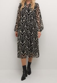 Robe à motifs noirs et beiges avec manches longues transparentes, détail avec nœud au col, silhouette fluide et ourlet mi-long. Associée à des bottines noires.