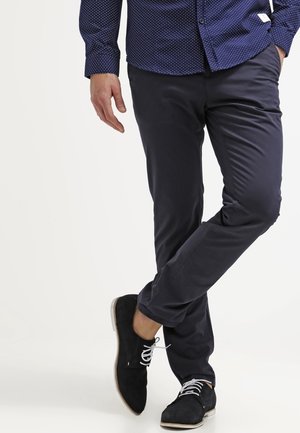 Marineblauwe slim-fit broek van lichte stof, met zijzakken en een gladde textuur, gecombineerd met zwarte suède schoenen en witte veters.