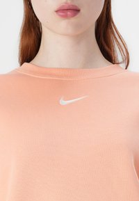 Sweatshirt pêssego feito de tecido macio, apresentando um decote canelado e um pequeno logo branco da Nike no peito. Textura suave sem padrões.