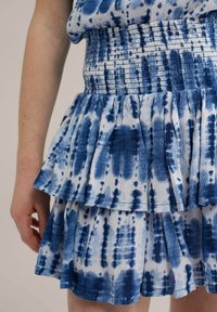 Fustă legată cu alb și albastru, cu talie elastică și volane suprapuse. Material moale și texturat cu un design fluid și un model aleator.