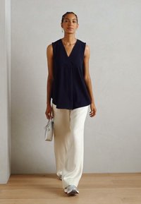 Blouse sans manches bleu marine avec décolleté en V, associée à un pantalon blanc fluide. Le modèle tient un sac à main argenté, affichant des détails minimaux et des textures lisses.