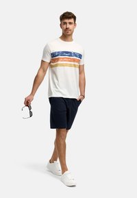 Hvid T-shirt med blå, orange og gule vandrette striber; marineblå shorts; hvide sneakers; holder solbriller; afslappet stil.