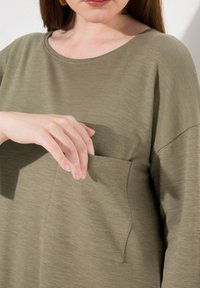 Olivgrünes, locker sitzendes Oberteil mit rundem Ausschnitt und kurzen Ärmeln; verfügt über eine Fronttasche und einen weichen, strukturierten Stoff.