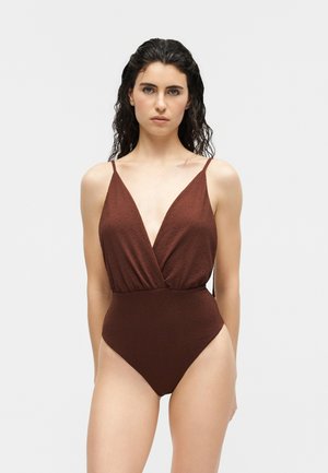 Femeie cu părul ud și întunecat, purtând un costum de baie maro texturat dintr-o singură piesă, cu bretele subțiri și un decolteu adânc în formă de V, stând în fața unui fundal simplu.
