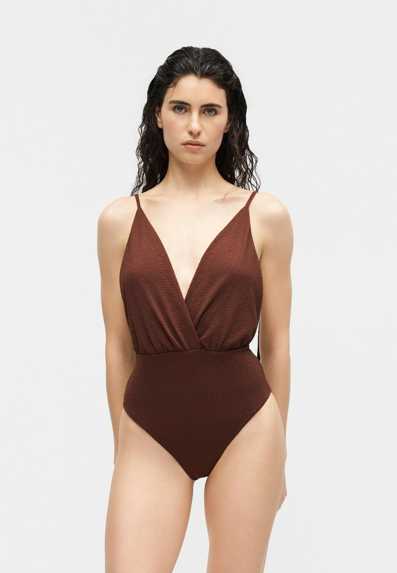 Femme aux cheveux foncés mouillés portant un maillot une pièce brun texturé avec de fines bretelles et un décolleté en V profond, debout devant un fond uni.