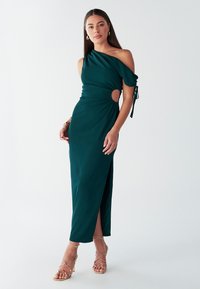 Abito monospalla turchese con un'apertura alla vita, tessuto arricciato e spacco alto sulla coscia. Abbinato a sandali dorati con cinturini.