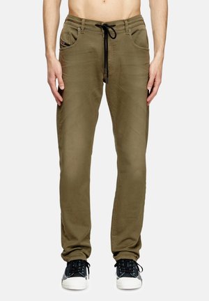 Homme portant un pantalon décontracté vert olive avec un cordon noir et des baskets noires et blanches, debout devant un fond clair uni.