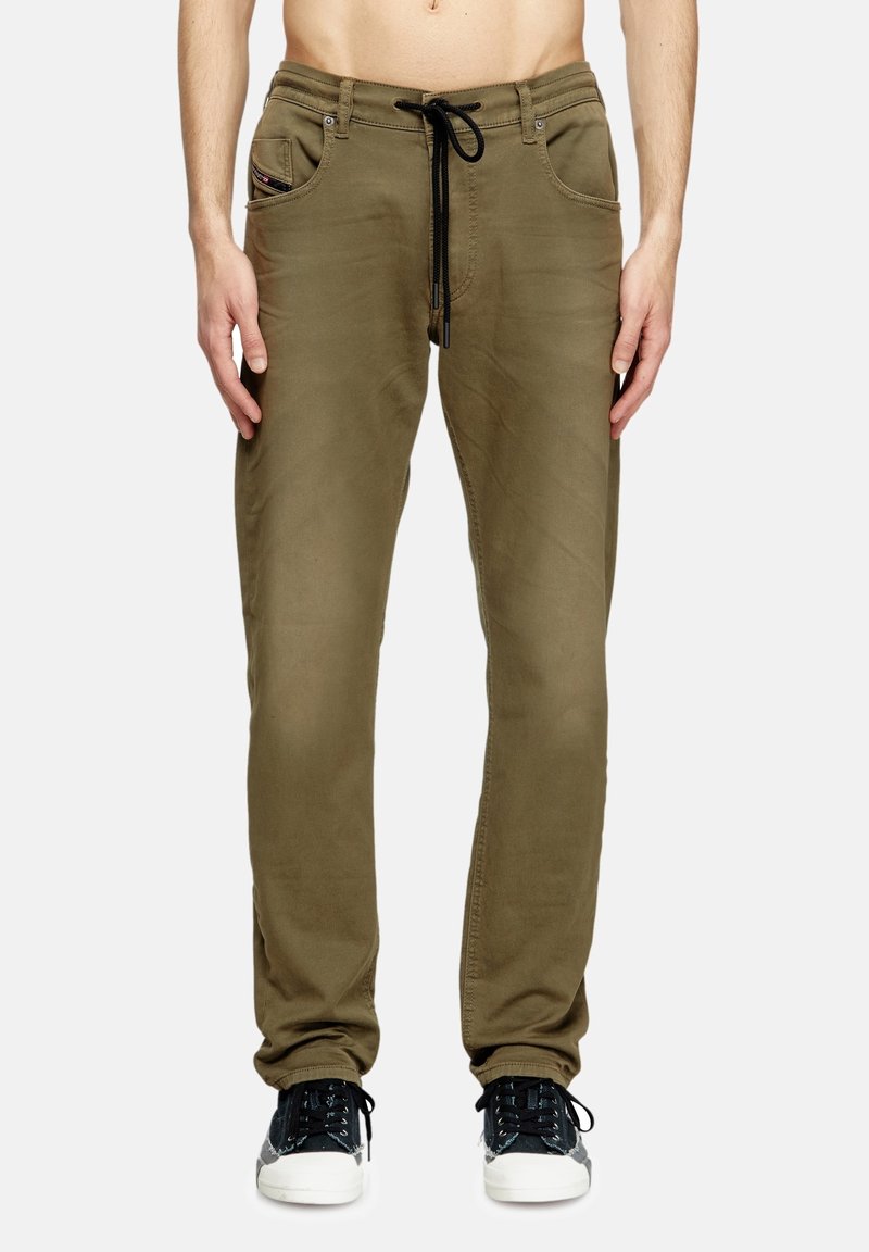 Homme portant un pantalon décontracté vert olive avec un cordon noir et des baskets noires et blanches, debout devant un fond clair uni.