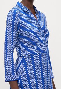Niebieska sukienka koszulowa z białym wzorem w zygzak, z zapinaniem na guziki z przodu, kołnierzykiem i podwiniętymi rękawami, wykonana z gładkiego materiału.
