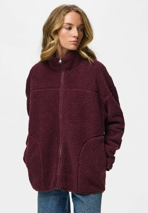 Jeune femme aux cheveux blonds ondulés portant une veste polaire bordeaux zippée et un jean bleu, regardant sur le côté sur fond blanc.