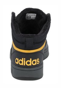 Zapatillas negras de media caña con un acabado texturado, detalles en amarillo, cuello acolchado y un destacado logo blanco de "adidas" en el talón.
