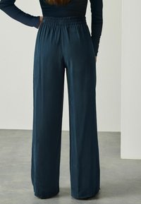 Vrouw in donkerblauwe hoge taille, wijde broek met een elastische tailleband, staand met één hand op haar heup tegen een effen achtergrond.