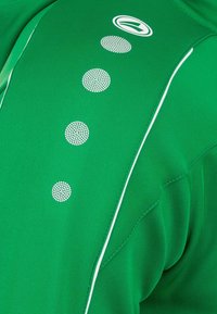 Maglietta atletica verde con una finitura testurizzata, caratterizzata da un logo e punti bianchi decorativi lungo la cucitura della spalla e cuciture a contrasto.