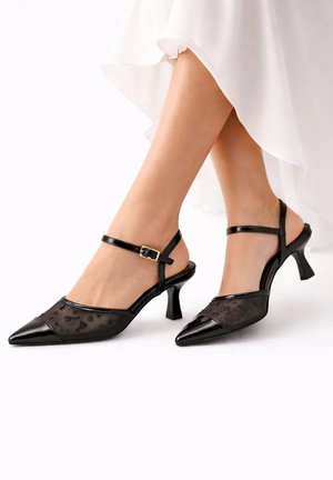 Frau trägt schwarze Slingback-Pumps mit spitzer Schuhspitze, Mesh und Blumendetails, kombiniert mit einem weißen, fließenden Rock.