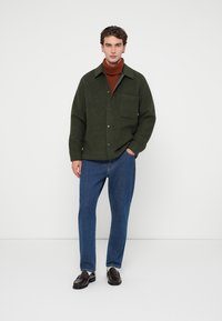 Nuori mies seisoo käsi taskussa, päällään vihreä napitettu takki, ruskea poolopaita, siniset farkut ja tummanruskeat loafersit.