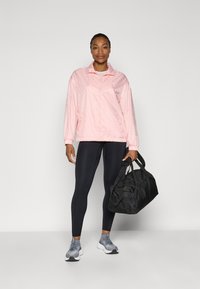 Lys rosa vindjakke med glidelås foran, elastiske ermer og en avslappet passform. Svart treningsbag og svarte tights kombinert med grå joggesko.