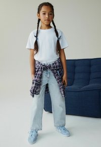 Tricou alb, blugi bleu deschis și adidași bleu deschis. O cămașă în carouri este legată în jurul taliei. Păr lung și întunecat împletit în împletituri.