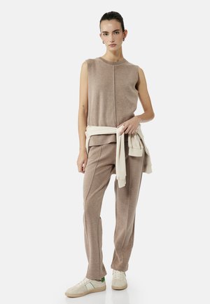 Vrouw staand, gekleed in een beige mouwloos topje en bijpassende broek, wit sweatshirt om de taille geknoopt en witte sneakers met groene accenten.