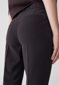 Pantalon sur mesure gris foncé avec une texture lisse, doté d'une taille plate et de poches arrière, présenté sous un angle latéral.