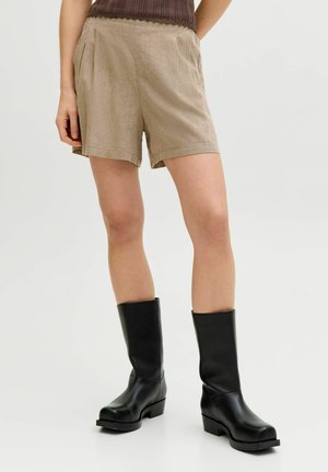 Személy bézs színű, magas derekú, pliszírozott rövidnadrágot, fekete középhosszú csizmát és barna bordázott felsőt visel egy egyszínű fehér háttér előtt.