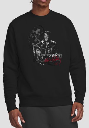 Mann trägt schwarzen Pullover mit schwarz-weißem Bild von Elvis Presley beim Gitarrenspielen, roter Signaturtext auf der Vorderseite.