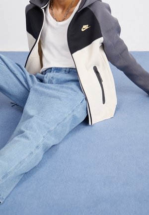 Osoba ubrana w czarno-biało-szarą kurtkę Nike zapinaną na zamek, białą koszulkę, srebrny naszyjnik łańcuszkowy oraz jasnoniebieskie dżinsy, siedząca na niebieskiej powierzchni.