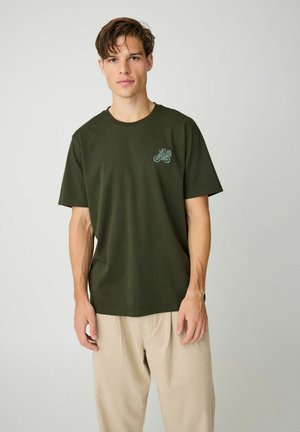 Ung mand iført mørkegrøn t-shirt med lille blå cykeldesign på brystet og beige bukser, står mod ensfarvet baggrund.