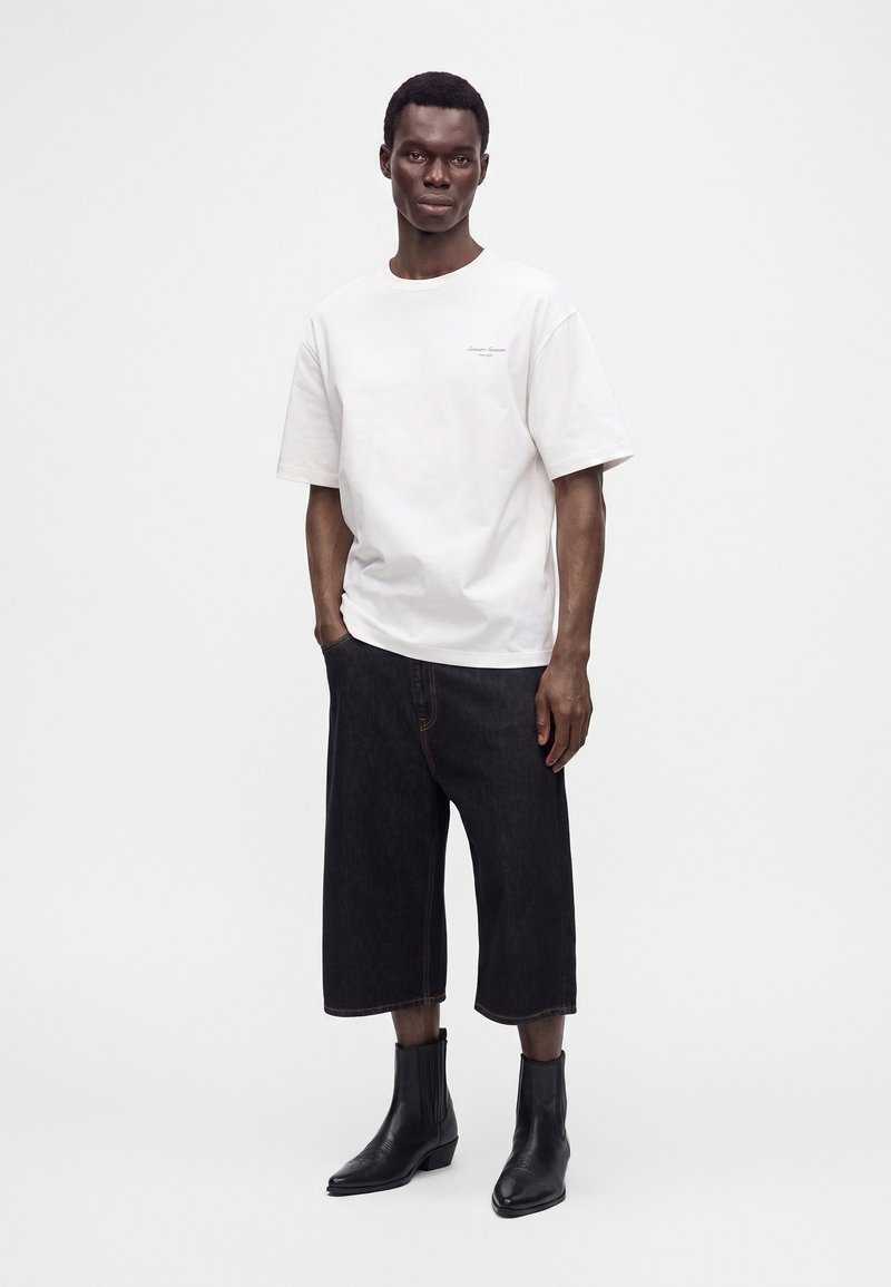 Homme debout portant un t-shirt blanc ample à manches courtes, un short noir large jusqu'aux genoux et des bottines noires sur un fond blanc uni.