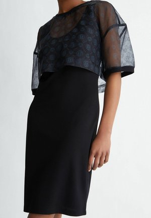 Kobieta w czarnej sukience bez rękawów, z przejrzystym, krótkim czarnym topem ozdobionym wzorami z okrągłymi logo oraz krótkimi rękawami.