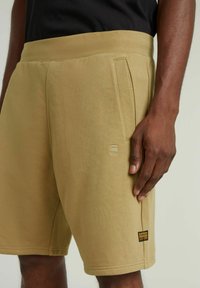 Pantaloni corte khaki con una vita ampia, tasche laterali e una toppa con logo. Realizzati in tessuto morbido con una texture liscia e cuciture pulite.