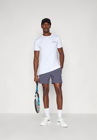 White short-sleeve t-shirt, gray athletic shorts, holding a tennis racket, wearing a black cap and white sneakers, in a neutral background.