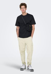 T-shirt en coton noir, pantalon cargo beige avec poches latérales, et baskets noires/blanches ; accessoirisé d'un petit sac bandoulière noir.