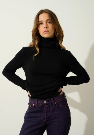 Musta kaulusneule, joka on valmistettu pehmeästä materiaalista, yhdistettyinä tumman violettiin farkkuun, jossa on kontrastivärinen tikkaus ja klassinen viiden taskun muotoilu.