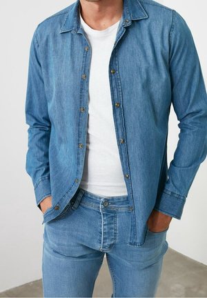 Man draagt lichtblauw spijkerhemd open over wit T-shirt en bijpassende lichtblauwe spijkerbroek, handen in de zakken.