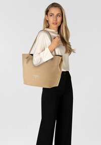 Bolso tote beige con textura suave, parte superior abierta y asas largas. Presenta un branding minimalista en la parte frontal. Combinado con una blusa de color neutro y pantalones negros.