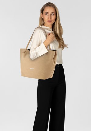 Beige schoudertas met een gladde textuur, open bovenkant en lange schouderbanden. Bevat minimale merkvermelding aan de voorkant. Gepaard met een top in neutrale kleur en zwarte broek.