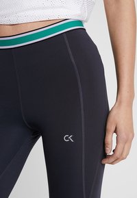 Close-up van een persoon die zwarte sportleggings draagt met een groen, wit en zwart elastiek en een klein "CK" logo op de dij.