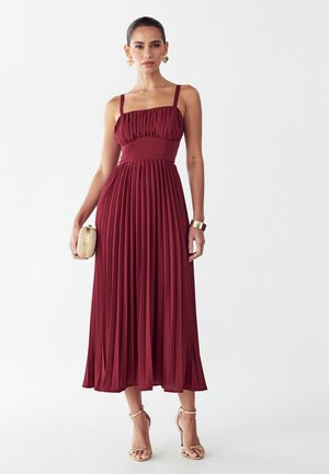 Vrouw draagt een mouwloze bordeaux plissé midi-jurk met een vierkante halslijn, houdt een beige clutch vast en draagt gouden sandalen en accessoires.