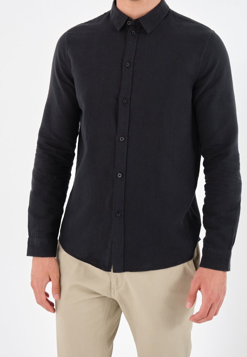 Camisa negra de manga larga hecha de tela texturada, con cuello abotonado y botones en la parte delantera, combinada con pantalones de color claro.