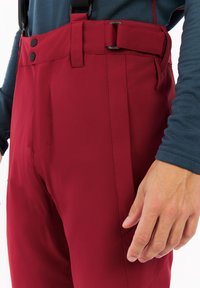 Rode outdoor broek gemaakt van zacht, rekbaar materiaal, met verstelbare bretels, knopen en een zijzak. Close-up weergave.