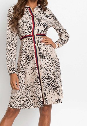 Donna che indossa un vestito beige al ginocchio con stampa animalier nera, bordo a righe rosse e blu navy, maniche lunghe e chiusura con bottoni sul davanti.